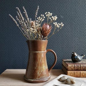 Peut inclure: Un pichet en céramique marron avec un bord évasé et une anse latérale, rempli d'un arrangement floral séché comprenant de la lavande, du gypsophile et une fleur de protéa. Le pichet présente un vernis terreux et tacheté aux nuances variées.