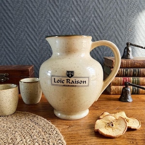 Puede incluir: Jarra de cerámica color crema con acabado moteado y asa grande. La jarra tiene el texto "Loïc Raison" en la parte delantera. Se ven dos tazas pequeñas a juego, junto con rodajas de manzana deshidratadas.