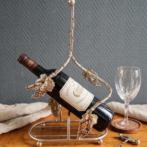 Puede incluir: Soporte para botella de vino de metal plateado con diseño de vid. Una botella de vino oscuro se sostiene en el centro, con una copa de vino y un sacacorchos cerca. La botella tiene una etiqueta que dice "Chateau Margaux".