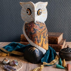 Peut inclure: Une figurine de hibou en bois sculpté avec une tête et un corps à finition craquelée blanche, dotée d'un motif d'aile style batik marron et noir. Le hibou a de grands yeux jaunes et noirs et repose sur une base noire, orné d'un tissu en velours bleu canard.