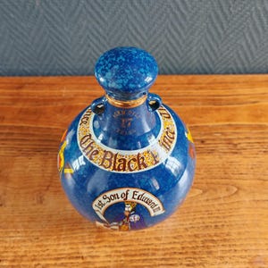 May include: A blue ceramic bottle with a matching stopper. The bottle has gold accents and text that reads "The Black Prince" and "1st Son of Edward". The bottle is aged over 17 years.