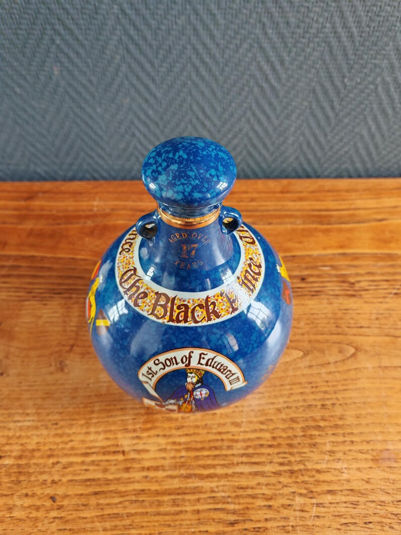May include: A blue ceramic bottle with a matching stopper. The bottle has gold accents and text that reads "The Black Prince" and "1st Son of Edward". The bottle is aged over 17 years.