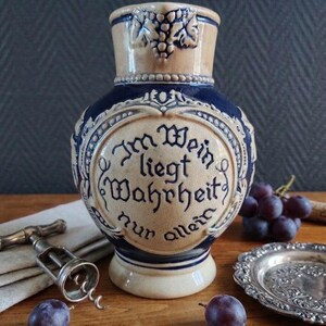 May include: A salt-glazed ceramic wine pitcher featuring a two-tone beige and cobalt blue finish. The front displays an embossed circular medallion with the German text Im Wein liegt Wahrheit nur allein, surrounded by intricate relief grape vine motifs.