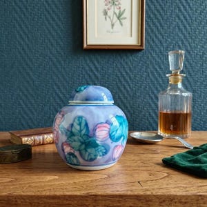 Peut inclure: Un pot en céramique décoratif avec un couvercle, orné de motifs floraux bleus, roses et verts. Une petite bouteille en verre et une cuillère sont également sur une surface en bois.