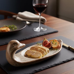 Peut inclure: Un plat de service en forme de canard contient des aliments tranchés, des figues et du pain grillé. Un verre de vin rouge et une assiette avec de la nourriture sont en arrière-plan. Le plat est sur une surface en ardoise noire.