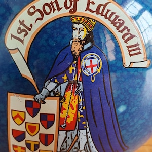 May include: A blue ceramic object with a historical illustration. The image features a crowned figure in a blue cloak, holding a shield with heraldic symbols. Text reads "1st Son of Edward III" and "of Chester".