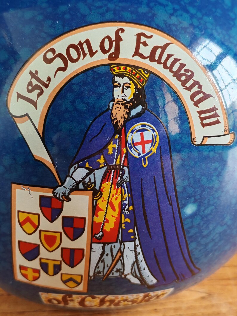 May include: A blue ceramic object with a historical illustration. The image features a crowned figure in a blue cloak, holding a shield with heraldic symbols. Text reads "1st Son of Edward III" and "of Chester".