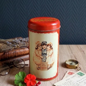 May include: A cylindrical red metal tin featuring a cream-colored label with an illustration of three children embracing. The label includes the text "WE DRINK Coca-Cola" and the Coca-Cola logo is printed on the top lid of the container.