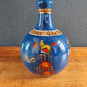 May include: A blue ceramic bottle with a decorative design. The bottle features a crown-shaped stopper with gold trim, and a label that reads "Prince The Royal". The bottle has a round body with a lion and flag design.