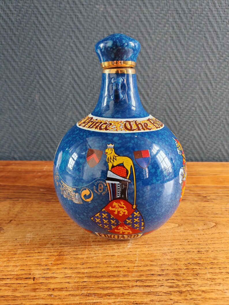 May include: A blue ceramic bottle with a decorative design. The bottle features a crown-shaped stopper with gold trim, and a label that reads "Prince The Royal". The bottle has a round body with a lion and flag design.