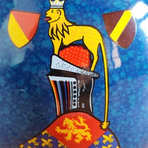 May include: A blue ceramic object with a yellow lion wearing a crown, red and yellow shields, and a red and gold crest. The lion is standing on a red and white base. The background is a textured blue.