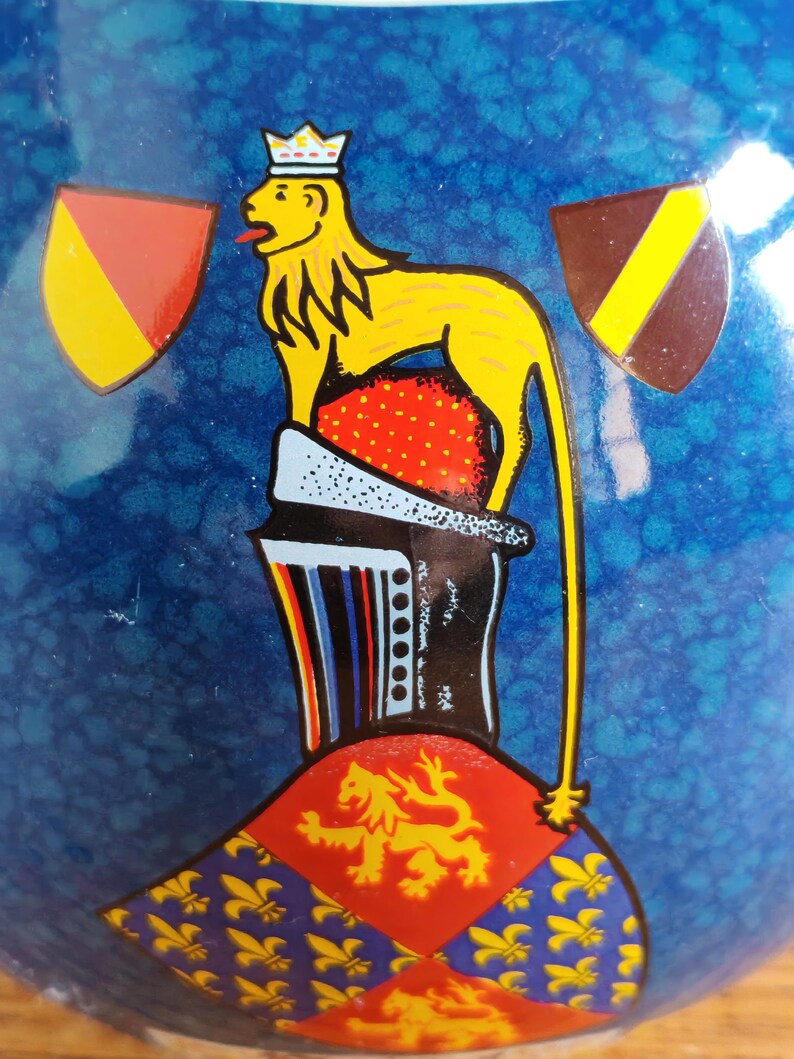 May include: A blue ceramic object with a yellow lion wearing a crown, red and yellow shields, and a red and gold crest. The lion is standing on a red and white base. The background is a textured blue.