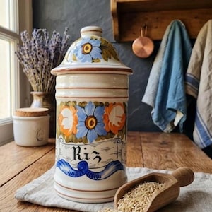 May include: A ceramic jar with a lid, decorated with blue and orange floral designs. The jar has the word "Riz" on it, and is sitting on a wooden table with a wooden scoop of rice.