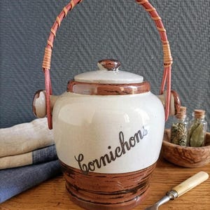 Puede incluir: Tarro de cerámica crema con tapa y asa trenzada. El tarro tiene detalles marrones y la palabra "Cornichons" en escritura cursiva. También se ven un tenedor vintage y pequeños frascos de vidrio.