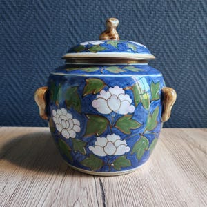 May include: A blue ceramic jar with a lid, featuring white floral designs and green leaves. The jar has two brown handles and a small brown figure on top of the lid. The jar is sitting on a wooden surface.