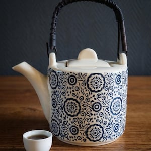 Peut inclure: Une théière en céramique couleur crème ornée d'un motif floral et de volutes bleu foncé, avec un couvercle assorti et une anse enveloppée de brun foncé. Une petite tasse en céramique blanche contenant un liquide sombre est posée à côté.