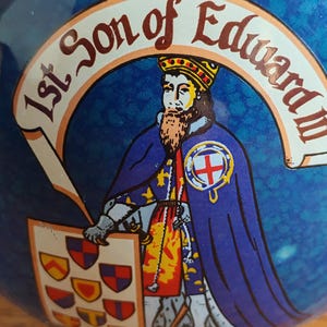 May include: A blue decorative item featuring a depiction of a historical figure. The figure wears a crown and a blue cape, holding a shield with crests. Text reads "1st Son of Edward III".