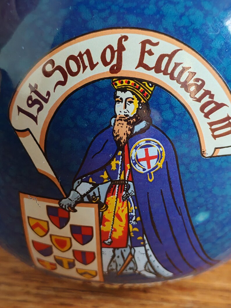 May include: A blue decorative item featuring a depiction of a historical figure. The figure wears a crown and a blue cape, holding a shield with crests. Text reads "1st Son of Edward III".