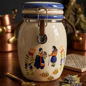 Op de afbeelding: Een crèmekleurige keramische pot met een blauw-wit deksel en een metalen sluiting. De pot is versierd met handgeschilderde figuren en bloemmotieven. Een gouden lepel en kruiden staan op de voorgrond.
