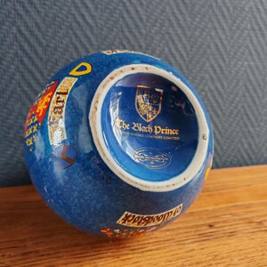 May include: A blue ceramic ashtray with a textured surface. The ashtray features a gold and red crest, the words "Earl" and "Woodstock", and the text "The Black Prince Scotch Whisky Company Limited".
