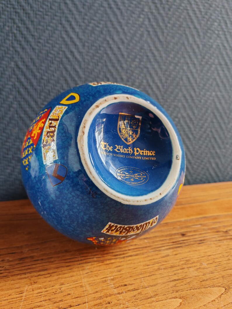 May include: A blue ceramic ashtray with a textured surface. The ashtray features a gold and red crest, the words "Earl" and "Woodstock", and the text "The Black Prince Scotch Whisky Company Limited".