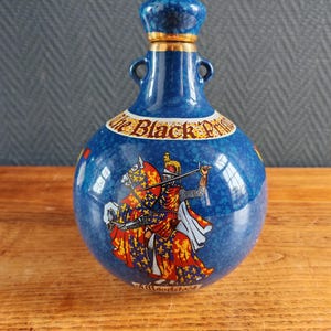 May include: A blue ceramic bottle with a decorative design. The bottle features a knight on horseback in red, yellow, and white. The text "The Black Prince" is visible on the bottle. The bottle has a blue stopper with gold trim.