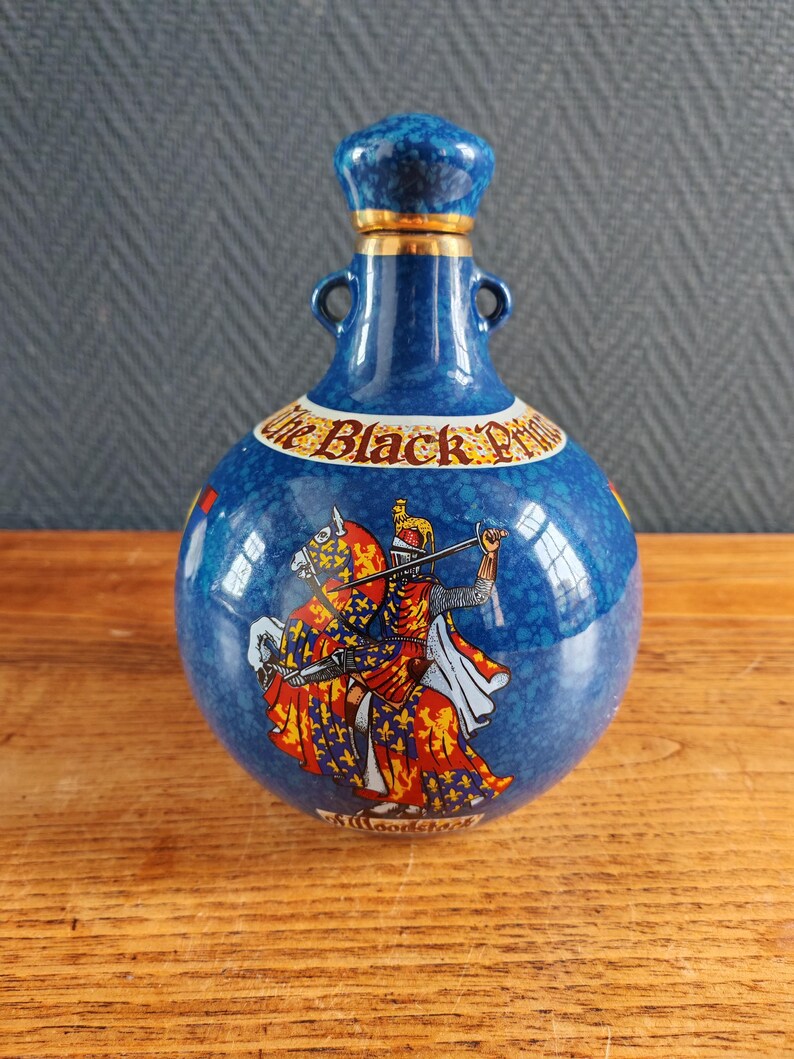 May include: A blue ceramic bottle with a decorative design. The bottle features a knight on horseback in red, yellow, and white. The text "The Black Prince" is visible on the bottle. The bottle has a blue stopper with gold trim.