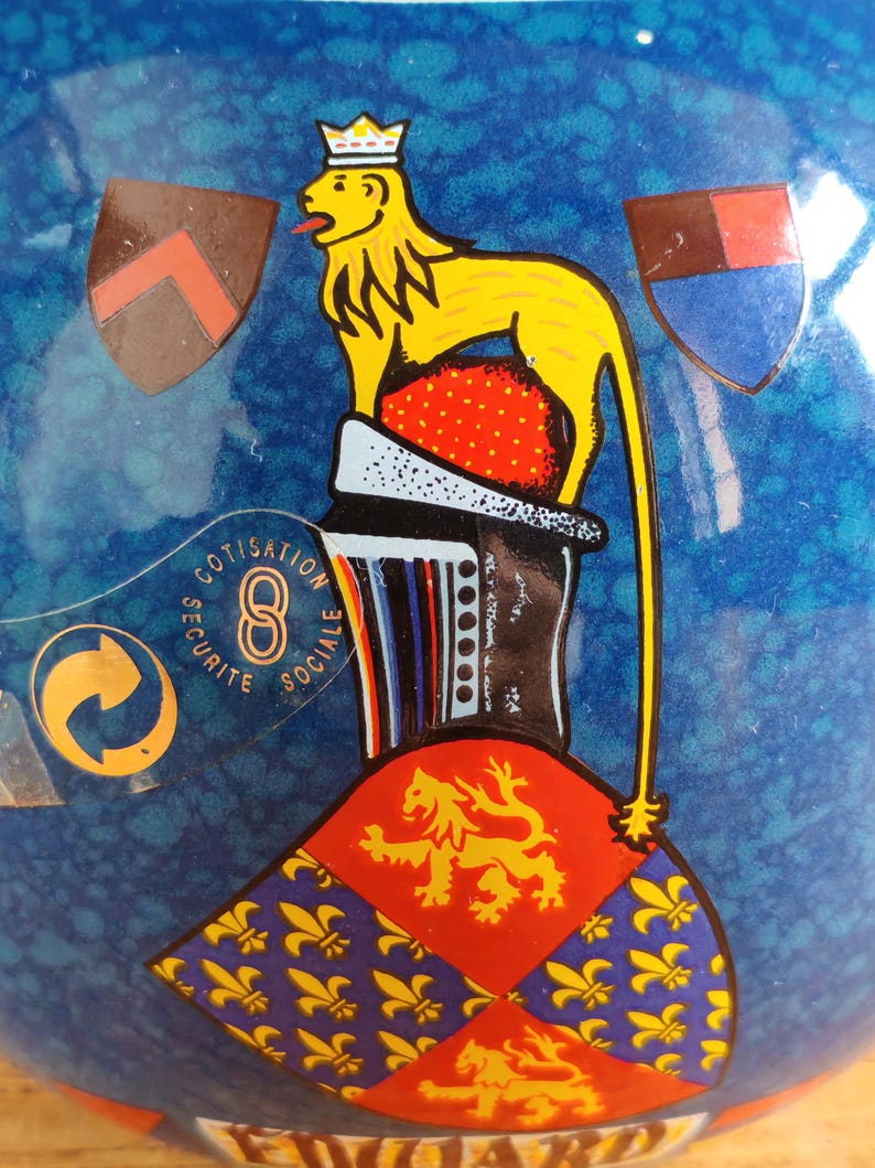 May include: A blue ceramic decorative object with a yellow lion and crown design. The object features a red and gold emblem, along with the text "COTISATION SECURITE SOCIALE".