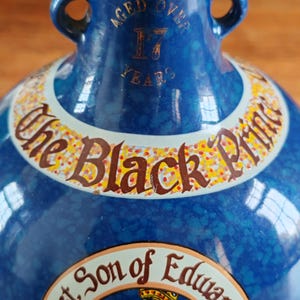 May include: A blue ceramic bottle with gold accents and two handles. The bottle features text that reads "Aged Over 17 Years" and "The Black Prince". A portrait of a man in a crown is also visible.