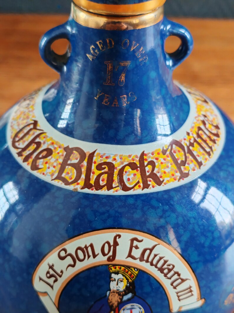 May include: A blue ceramic bottle with gold accents and two handles. The bottle features text that reads "Aged Over 17 Years" and "The Black Prince". A portrait of a man in a crown is also visible.