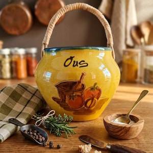 Puede incluir: Una olla de cerámica amarilla con asa trenzada, con una ilustración pintada de verduras y la palabra "Jus". La olla está sobre una superficie de madera con especias, hierbas y un cuenco de madera.