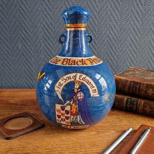 May include: A blue ceramic bottle with a decorative design. The bottle features a portrait of a king and the text "1st Son of Edward III". The bottle has a blue stopper and two small handles. The bottle is sitting on a wooden surface.