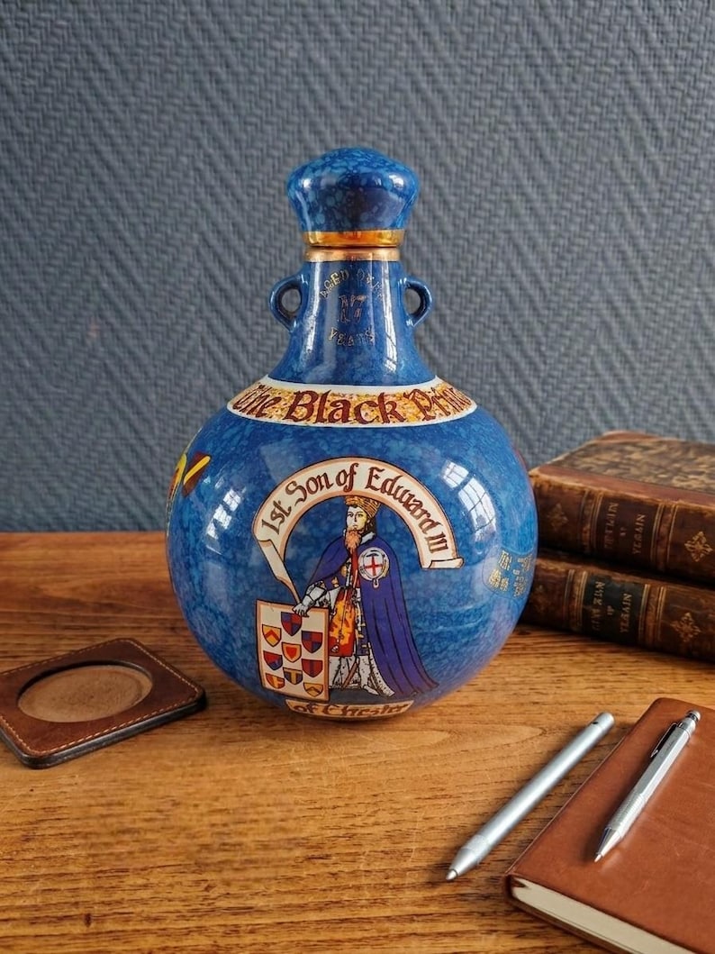 May include: A blue ceramic bottle with a decorative design. The bottle features a portrait of a king and the text "1st Son of Edward III". The bottle has a blue stopper and two small handles. The bottle is sitting on a wooden surface.