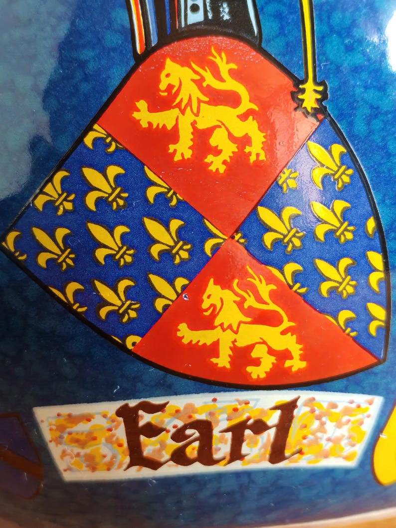 May include: Close-up of a shield design with a blue background. The shield is divided into sections of red with a yellow lion and blue with yellow fleur-de-lis. The word "Earl" is written in brown script.