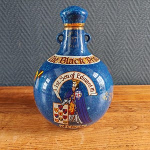 May include: A blue ceramic bottle with a matching stopper. The bottle features a gold rim and handles, with text and an illustration of a figure in royal attire. The text reads "The Black Prince" and "1st Son of Edward III".