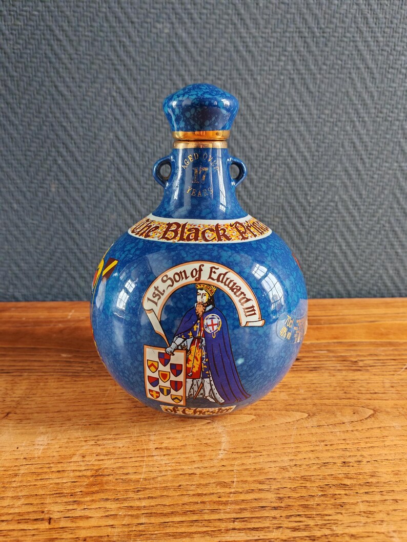 May include: A blue ceramic bottle with a matching stopper. The bottle features a gold rim and handles, with text and an illustration of a figure in royal attire. The text reads "The Black Prince" and "1st Son of Edward III".