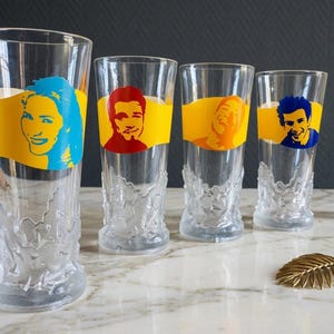 May include: Four clear drinking glasses with yellow bands featuring colourful portraits. The glasses have a textured base and are arranged on a marble surface. A gold leaf and perfume bottle are also present.