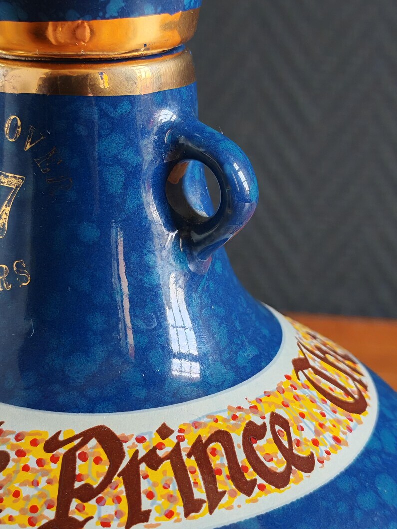 May include: Close-up of a blue ceramic bottle with gold accents. The bottle has a handle and a decorative band with text. The bottle's surface has a speckled pattern. The text on the bottle reads "OVER 17 YEARS" and "Prince".