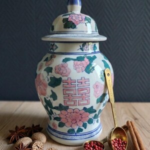 May include: A white ceramic ginger jar with a lid, decorated with pink floral patterns, green leaves, and a double happiness symbol in pink. Beside it are brass measuring spoons containing pink peppercorns, star anise, nutmeg, and cinnamon sticks.