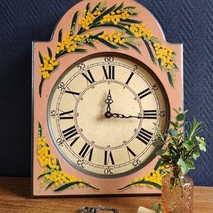 Peut inclure: Une horloge murale de style vintage avec un cadre rose clair et un cadran crème. L'horloge présente des chiffres romains et des aiguilles noirs, avec des motifs floraux jaunes. Une clé décorative et un petit vase avec de la verdure sont devant l'horloge.