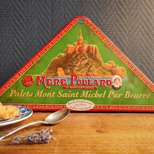 Puede incluir: Una lata triangular de galletas "Palets Mont Saint Michel Pur Beurre", con un diseño verde y rojo y una ilustración del Mont Saint Michel. Un plato de galletas, una cuchara y ramitas de lavanda están en primer plano.