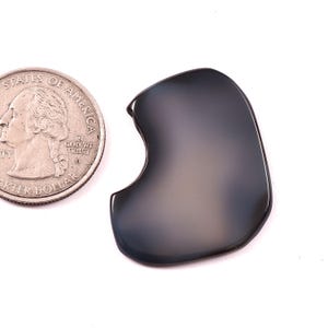May include: A smooth, dark grey, irregularly shaped stone cabochon with a polished finish, positioned next to a United States quarter coin for scale. The stone has a curved, organic silhouette with a distinct indentation on one side.