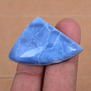 Peut inclure: Une pierre précieuse triangulaire polie, de couleur bleu clair, avec un réseau de veines blanches. La pierre a une surface lisse et brillante et est tenue entre deux doigts. La forme générale rappelle une part de tarte.