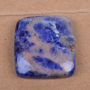 Puede incluir: Una gema de sodalita de forma cuadrada con esquinas redondeadas. La piedra presenta un color azul profundo con vetas blancas y rosas. La superficie pulida refleja la luz, resaltando los patrones y texturas naturales de la piedra.