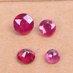 May include: Four faceted, round-cut rubies in varying sizes. The gemstones display a deep red hue with reflective facets, showcasing their clarity and brilliance. The rubies are set against a neutral background, highlighting their color and shape.