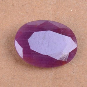 Op de afbeelding: Ovaal geslepen robijn edelsteen met een diepe rood-paarse tint. Het gefacetteerde oppervlak reflecteert licht, wat de helderheid en schittering van de edelsteen benadrukt. De edelsteen ligt op een neutrale achtergrond.
