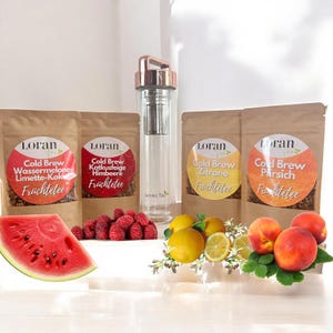 Op de afbeelding: Een heldere glazen waterfles met een roségouden deksel en infuser staat tussen vier zakken cold brew thee, een watermeloen schijf, frambozen, citroenen en perziken. De theesmaken zijn watermeloen, framboos, citroen en perzik.