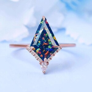 May include: A rose gold ring featuring a kite-shaped, dark blue gemstone with colorful inclusions. The gemstone is set with small, clear stones and a pear-shaped accent. The ring is set against a light blue background.