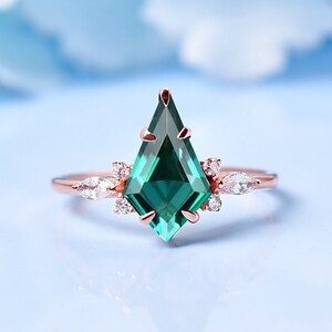 May include: A rose gold ring with a large, emerald green, kite-shaped gemstone surrounded by smaller, clear gemstones.