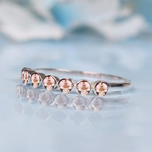 May include: A silver ring with seven small, round, peach-colored gemstones set in a row. The ring is displayed on a reflective, light blue surface, with a blurred, light blue and white background.
